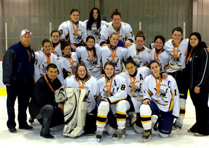 A photo of a women's hockey team and their coaches. They are posed in 3 rows on the ice of a skating rink. In the front row, there are 4 women kneeling, one of them being Chelsea with a Captain badge on her jersey. At the end of the front row and both ends of the second row are coaches wearing black jackets and pants. The team members and their coaches are all wearing medals around their neck. The ribbon of the metals is orange. 