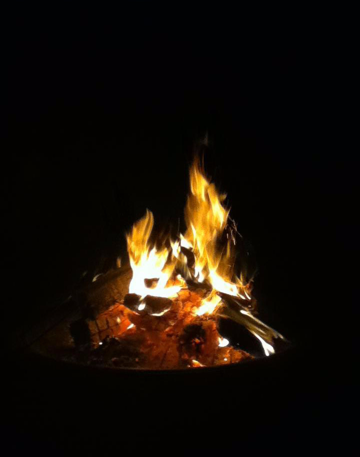 A photo of a bonfire pit a night with a fire lit in the centre. The background is all black and the pit is lit up by the fire. There is red burning embers underneath the fire.