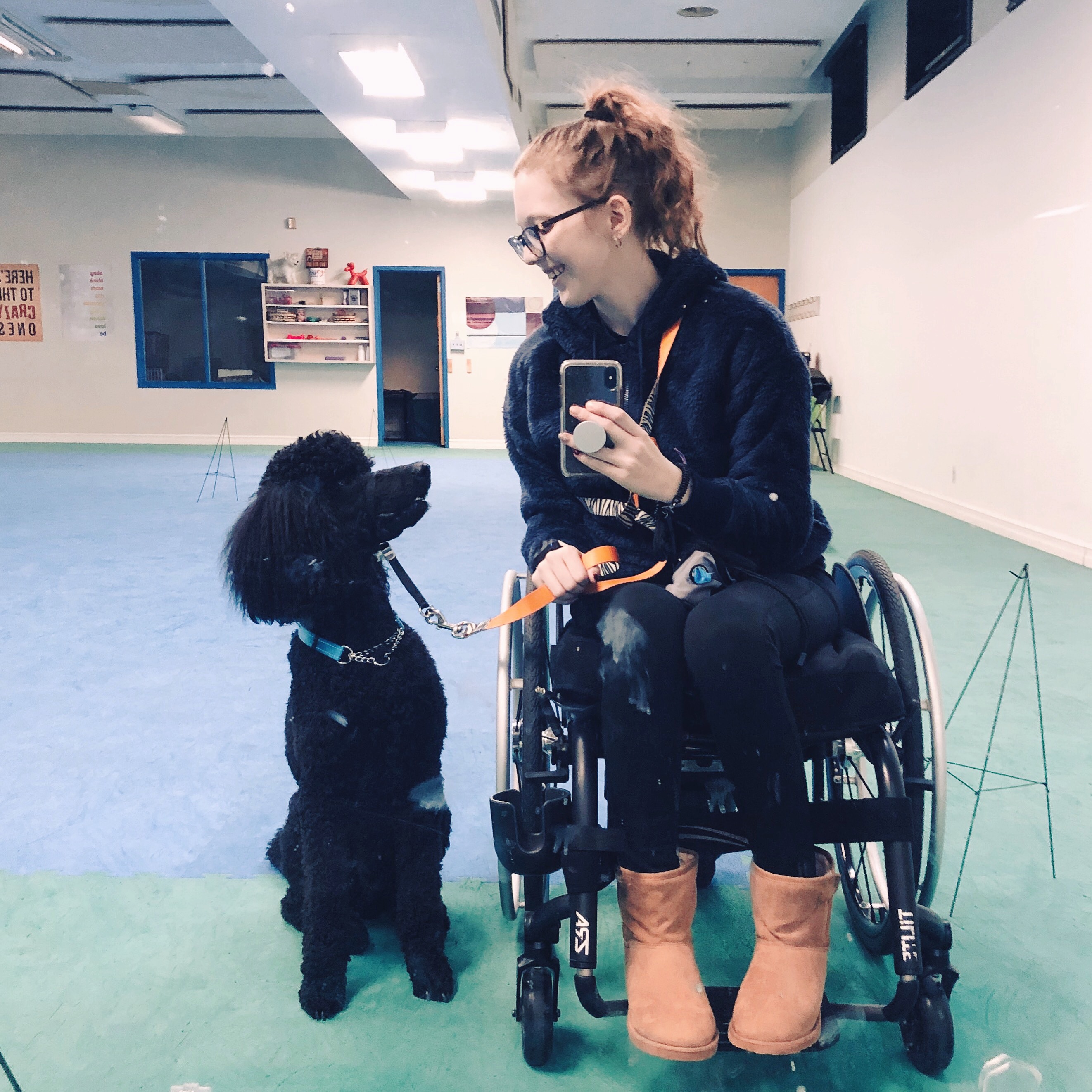 A photo of Kathleen siting in a small black wheelchair at the dog gym. She is leaning to the left towards her black poodle who is seated beside her. The dog is look up at Kathleen and she is looking down at him, smiling. She is holding her phone up at chest height using it to take a mirror selfie. Kathleen is being glasses and has wavy dirty blonde hair pulled back into a ponytail. She is wearing a navy hoodie, black leggings, and tan ugg style boots. In the background, you can see the floor covered in blue mats with green mats around the periphery. The wall behind them is white and has a open door and window as well as a bookshelf. 